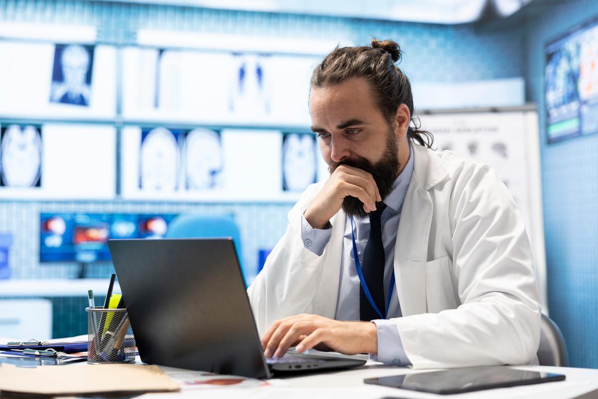Doctor reviewing records on a laptop with X-rays displayed on the wall behind.