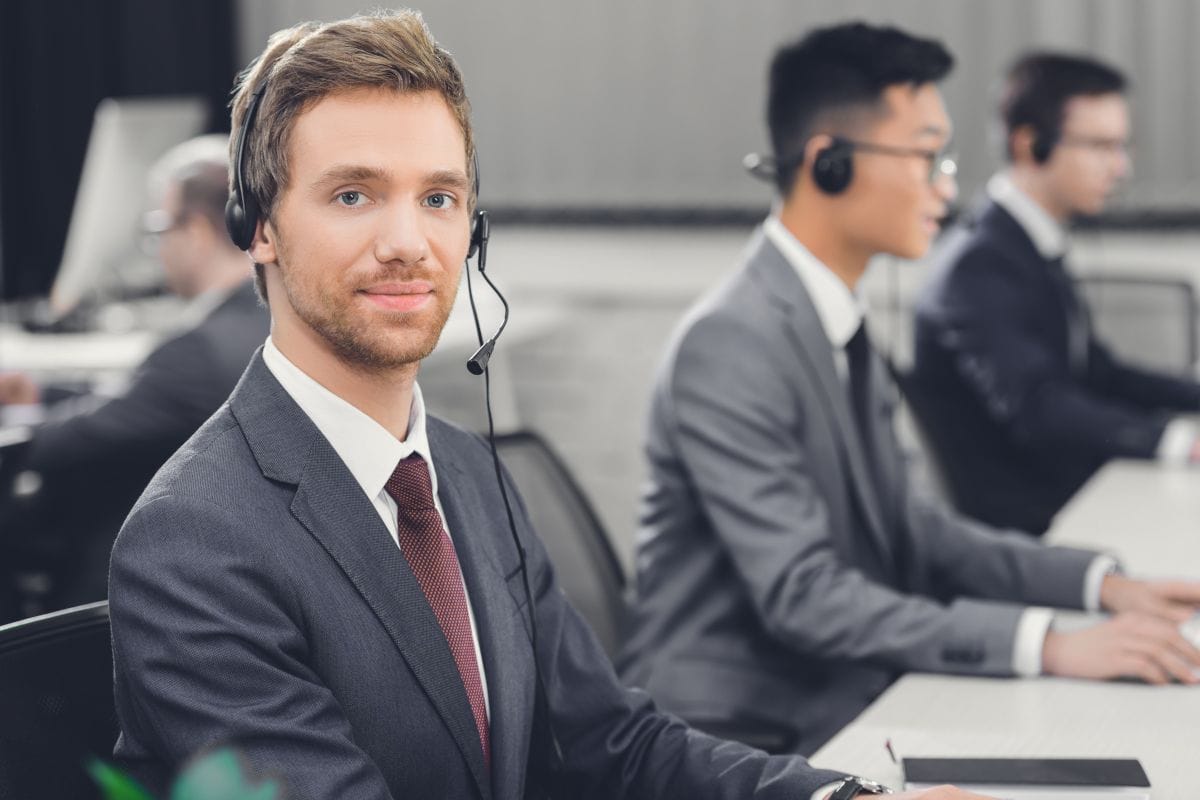 Smiling helpdesk technician in a headset at his desk, backed by a team of IT experts managing client systems every day.