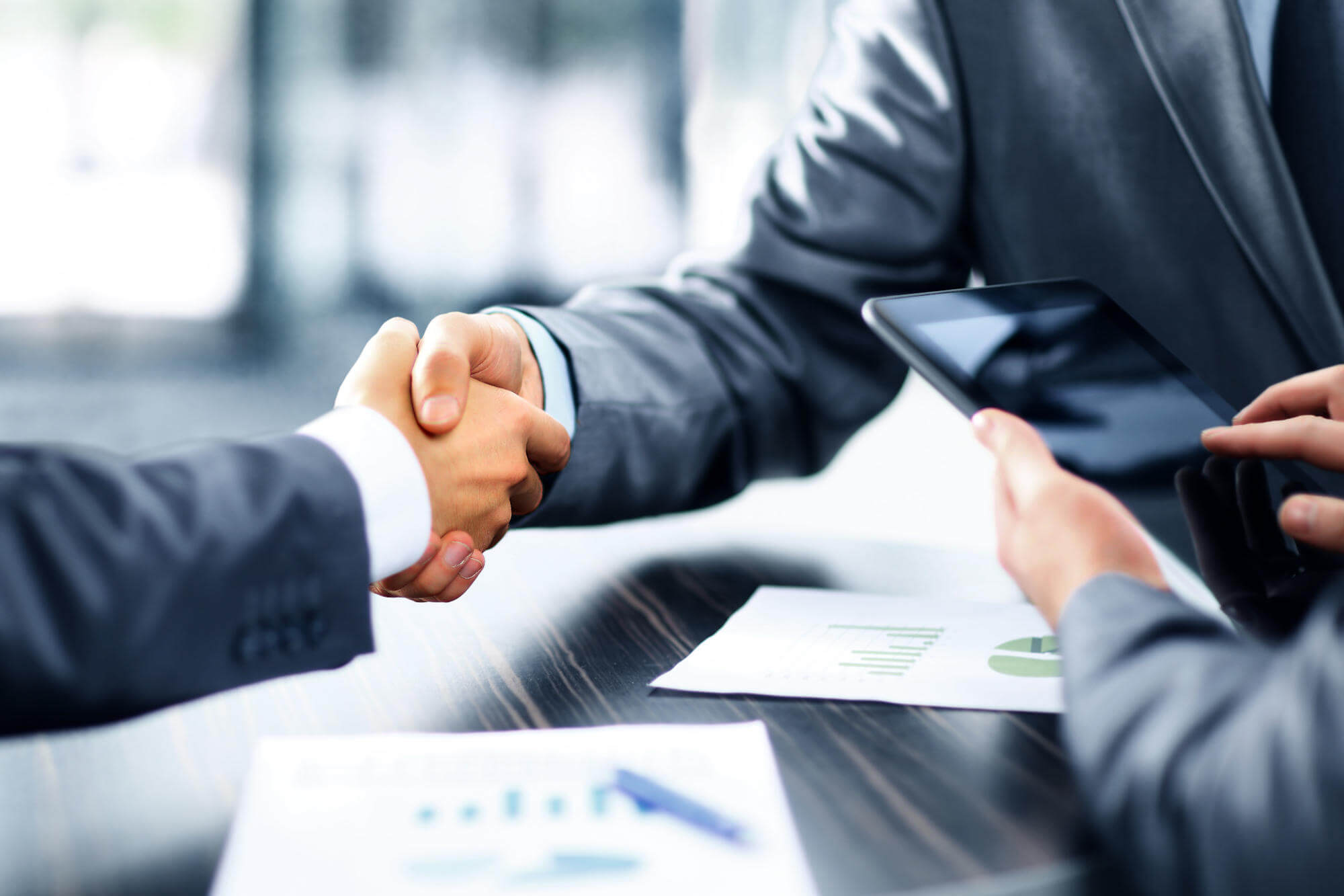 Two business people shaking hands over a conference table with a tablet and printed charts after agreeing on IT services